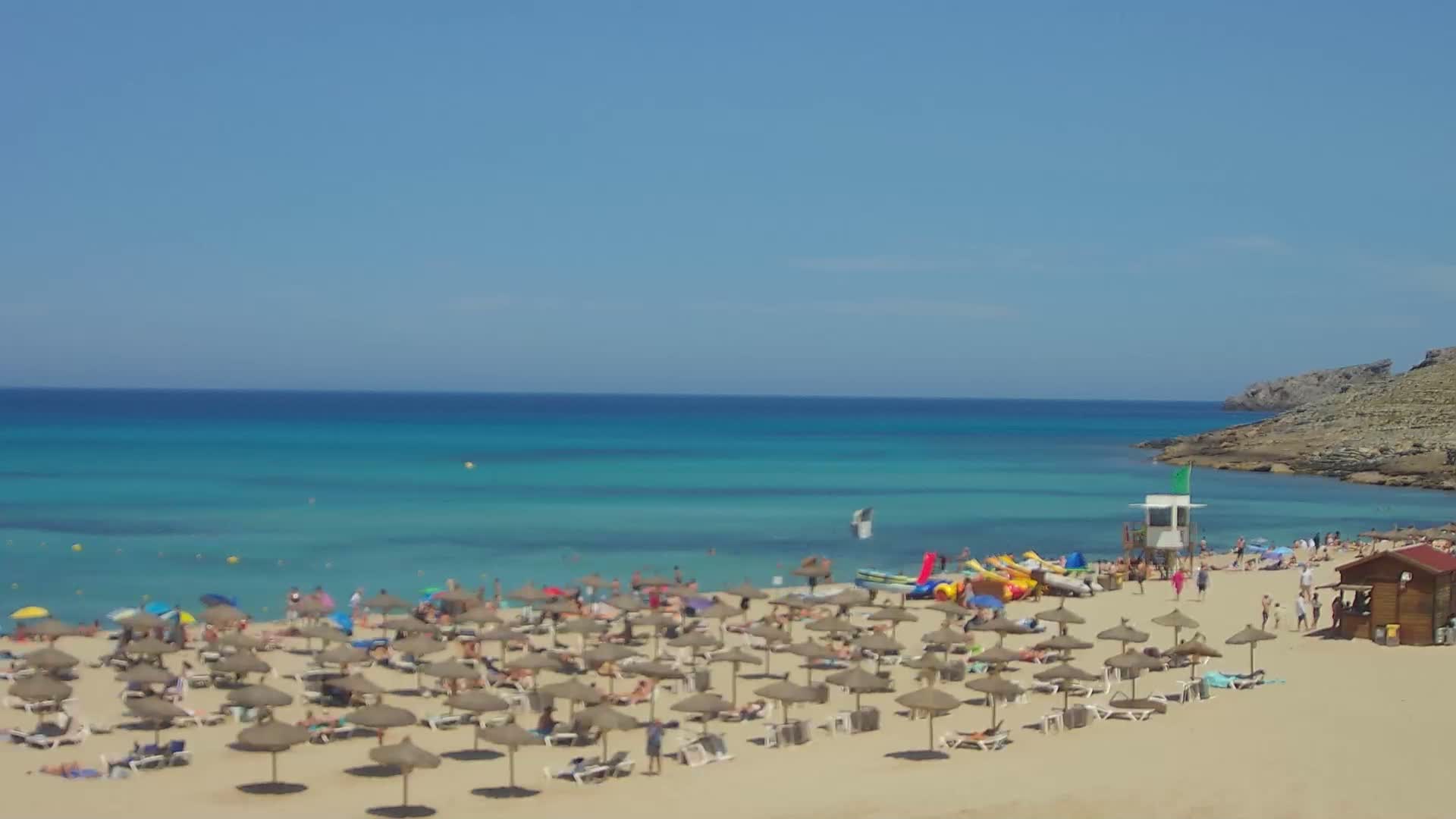 A sandy beach with numerous straw umbrellas and beachgoers is nestled next to a calm, turquoise ocean on a sunny day.