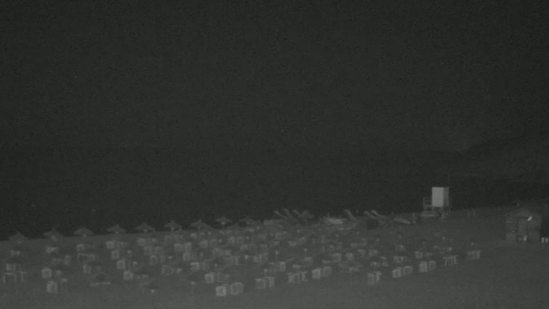 A nighttime beach scene shows numerous beach umbrellas and chairs under a dark sky.