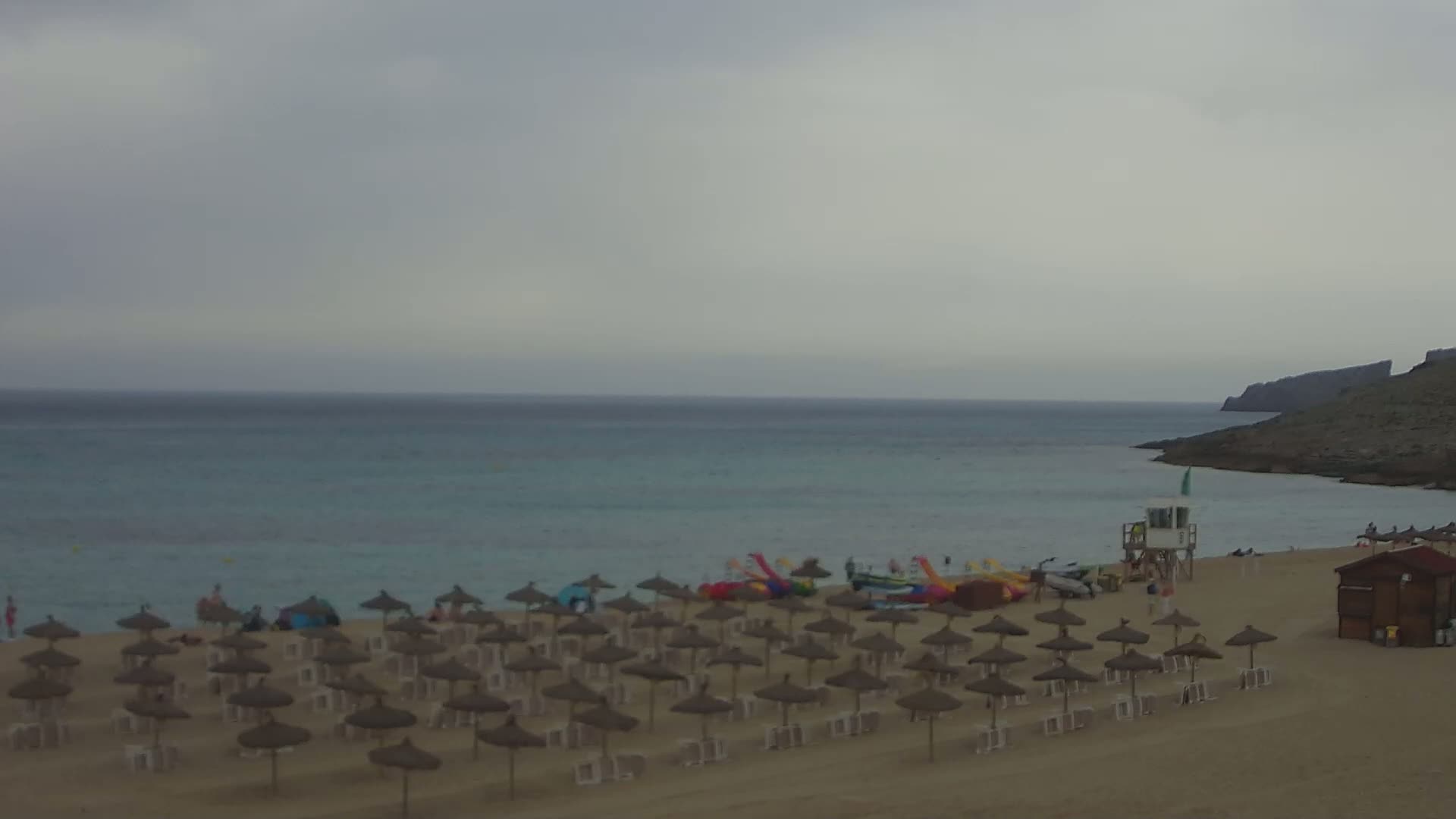 A sandy beach with numerous straw umbrellas under an overcast sky overlooks a calm, turquoise sea and a rocky headland.