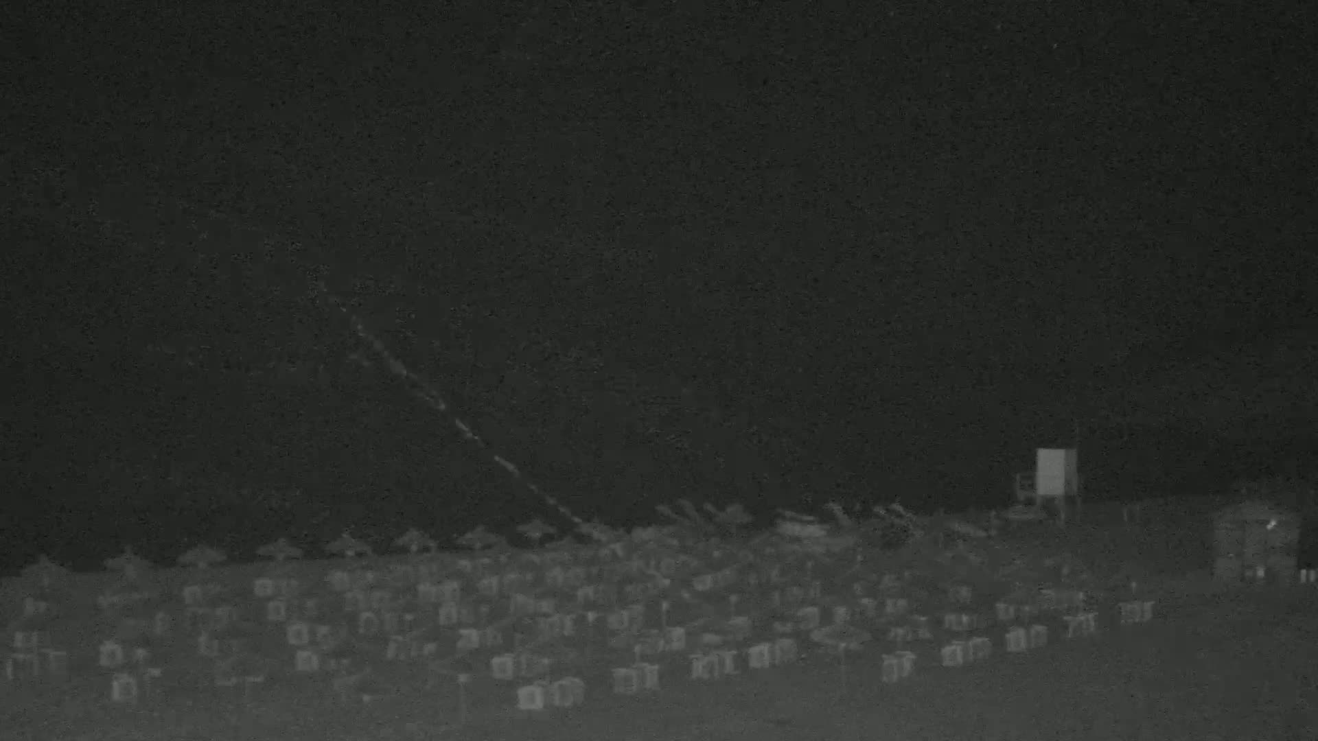 Under a dark night sky, a large number of objects, possibly beach chairs, are arranged on a flat surface, with a few structures visible in the distance.