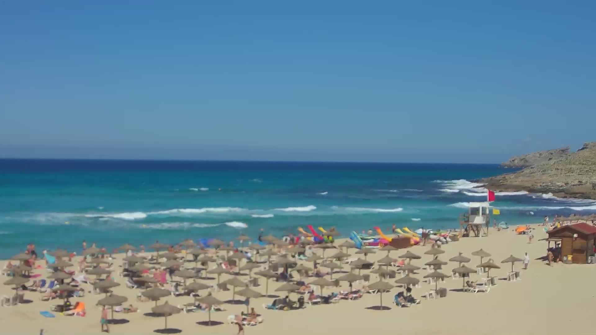 A sandy beach with many thatched umbrellas and people enjoying a sunny day with turquoise water and small waves.
