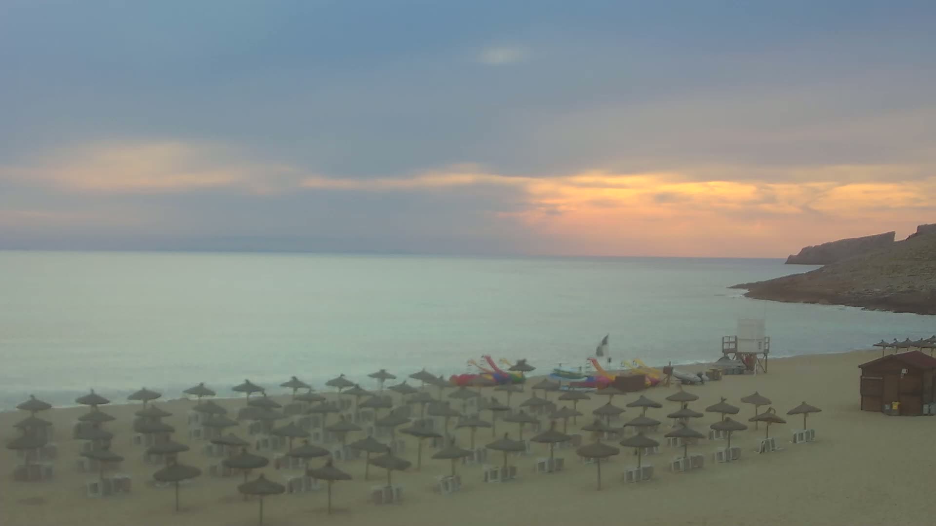 A sandy beach with numerous thatched umbrellas under a pale sunset sky over calm, light-green water, backed by a rocky cliff.