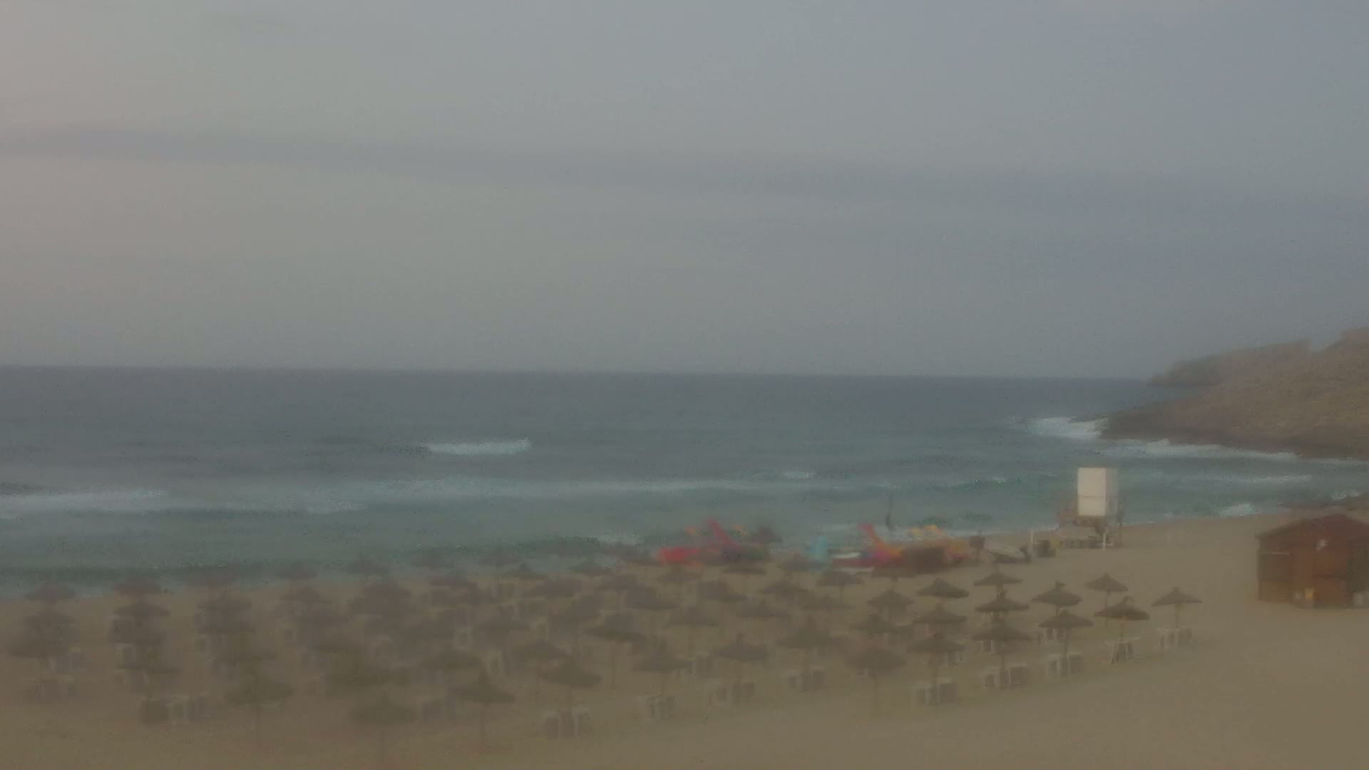 A hazy, overcast day at a beach shows a sandy shore with numerous closed umbrellas and a lifeguard stand, and gentle waves lapping at the shoreline.