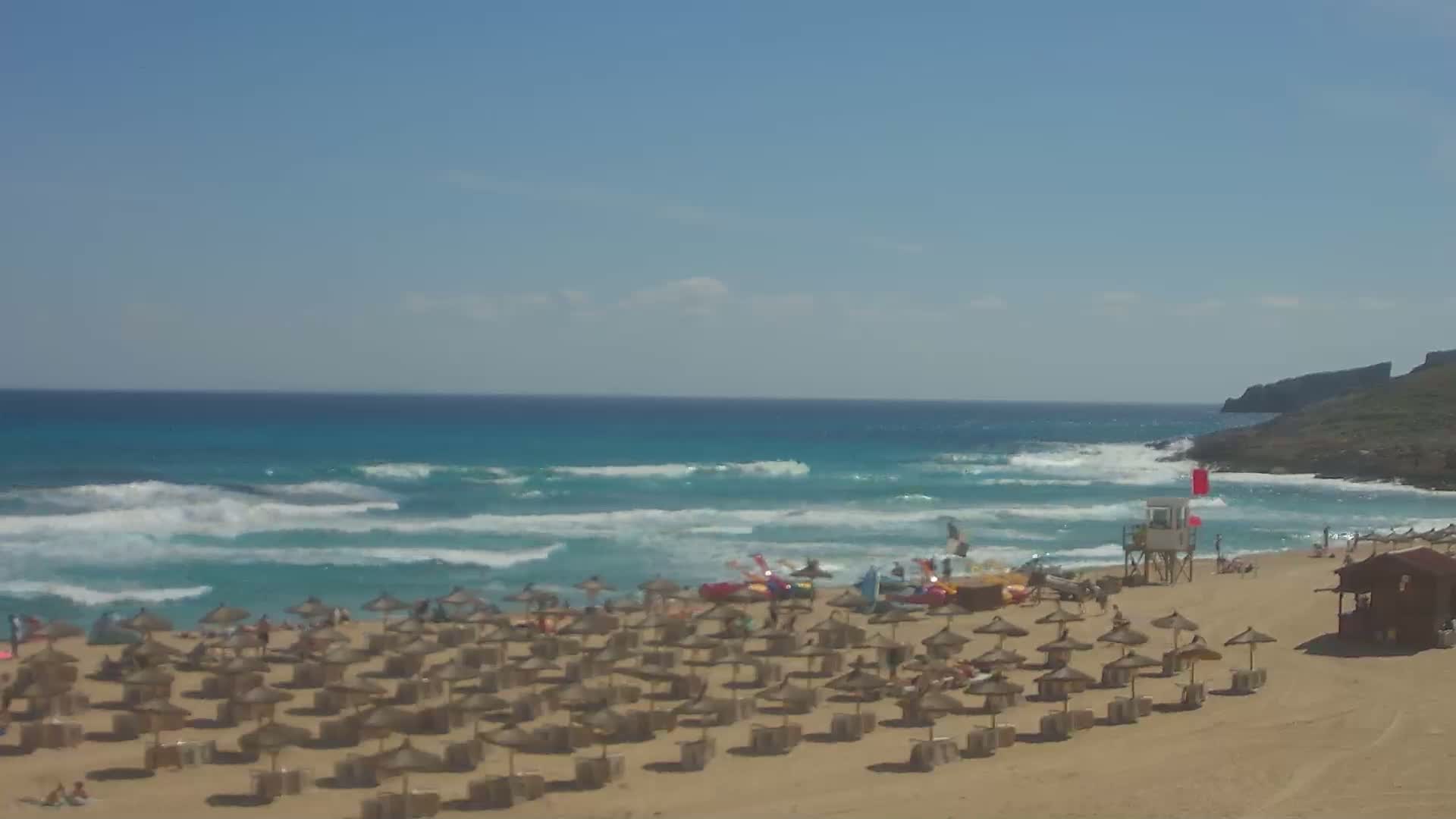 A sandy beach with numerous straw umbrellas, a lifeguard tower, and several people is shown under sunny skies, with moderate waves rolling into the turquoise ocean.