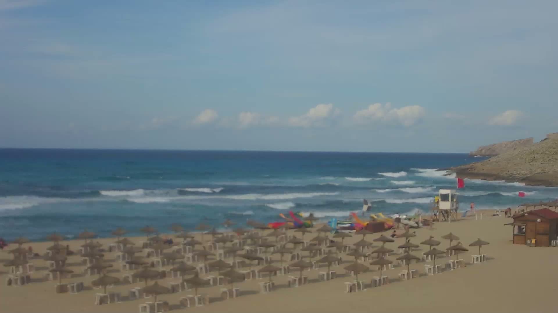 A sandy beach with numerous straw umbrellas, a lifeguard stand, and a small building is situated beside a turquoise ocean with small waves under a partly cloudy sky.