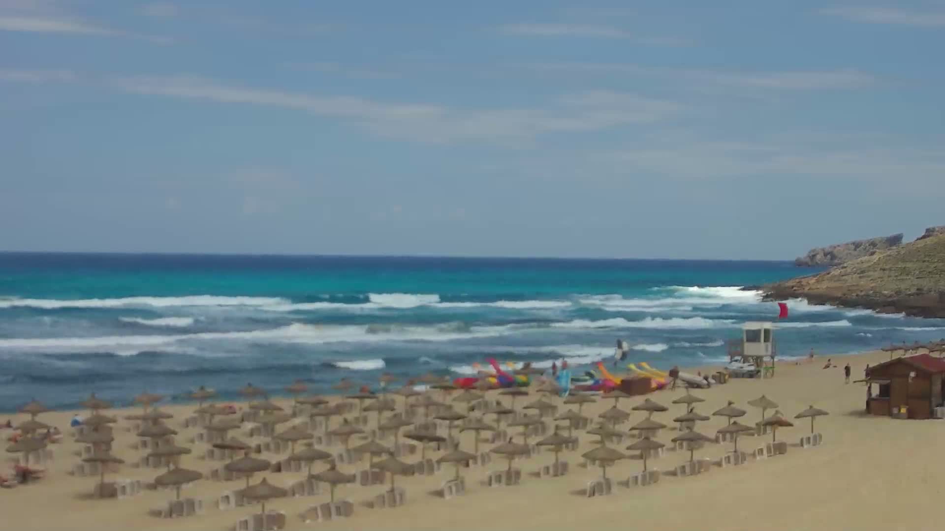 A sandy beach with numerous straw umbrellas and a lifeguard stand is situated before a turquoise ocean with moderate waves under a partly cloudy sky.