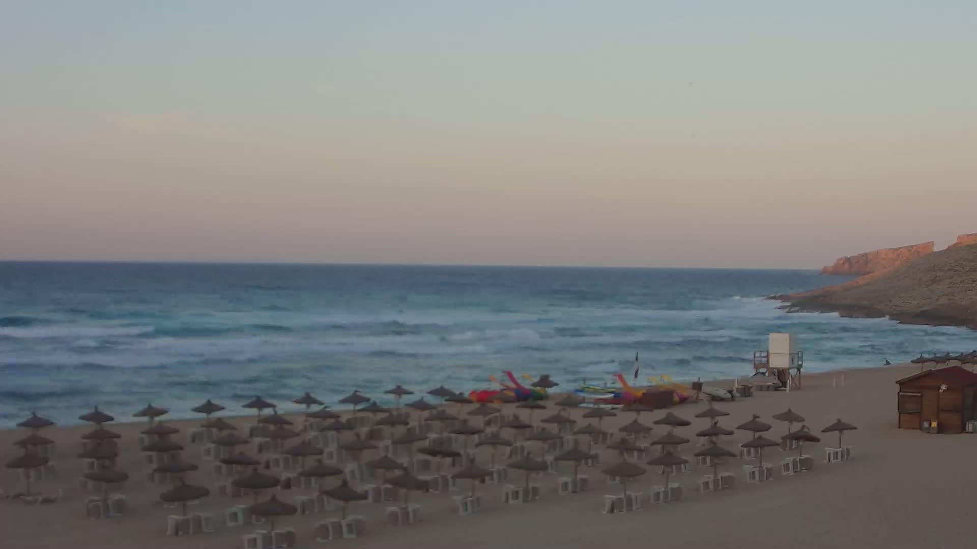 Many straw umbrellas line a sandy beach next to a calm, slightly wavy ocean under a pale, sunset sky, with a lifeguard stand and a small building visible.