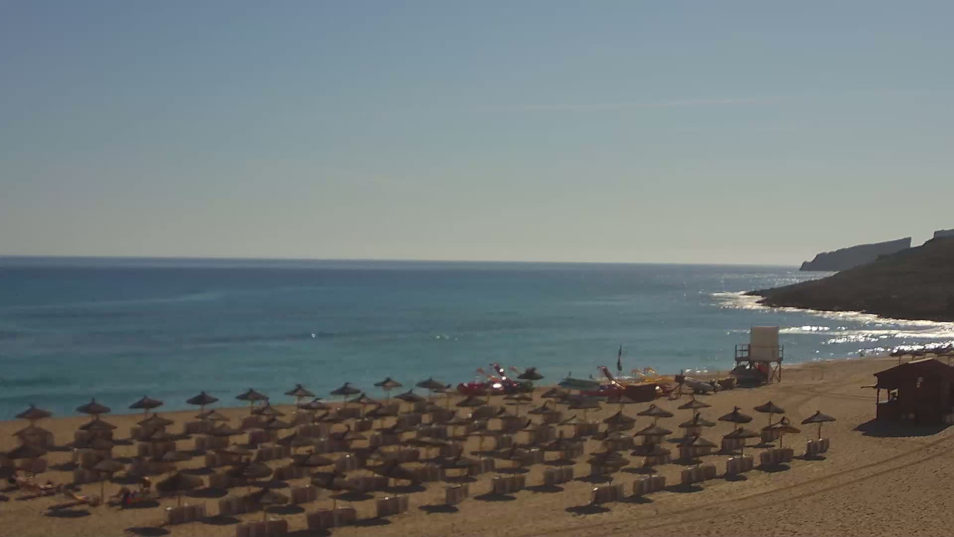 A sandy beach with numerous straw umbrellas, a lifeguard stand, and small boats sits on a calm, clear ocean under a sunny sky.