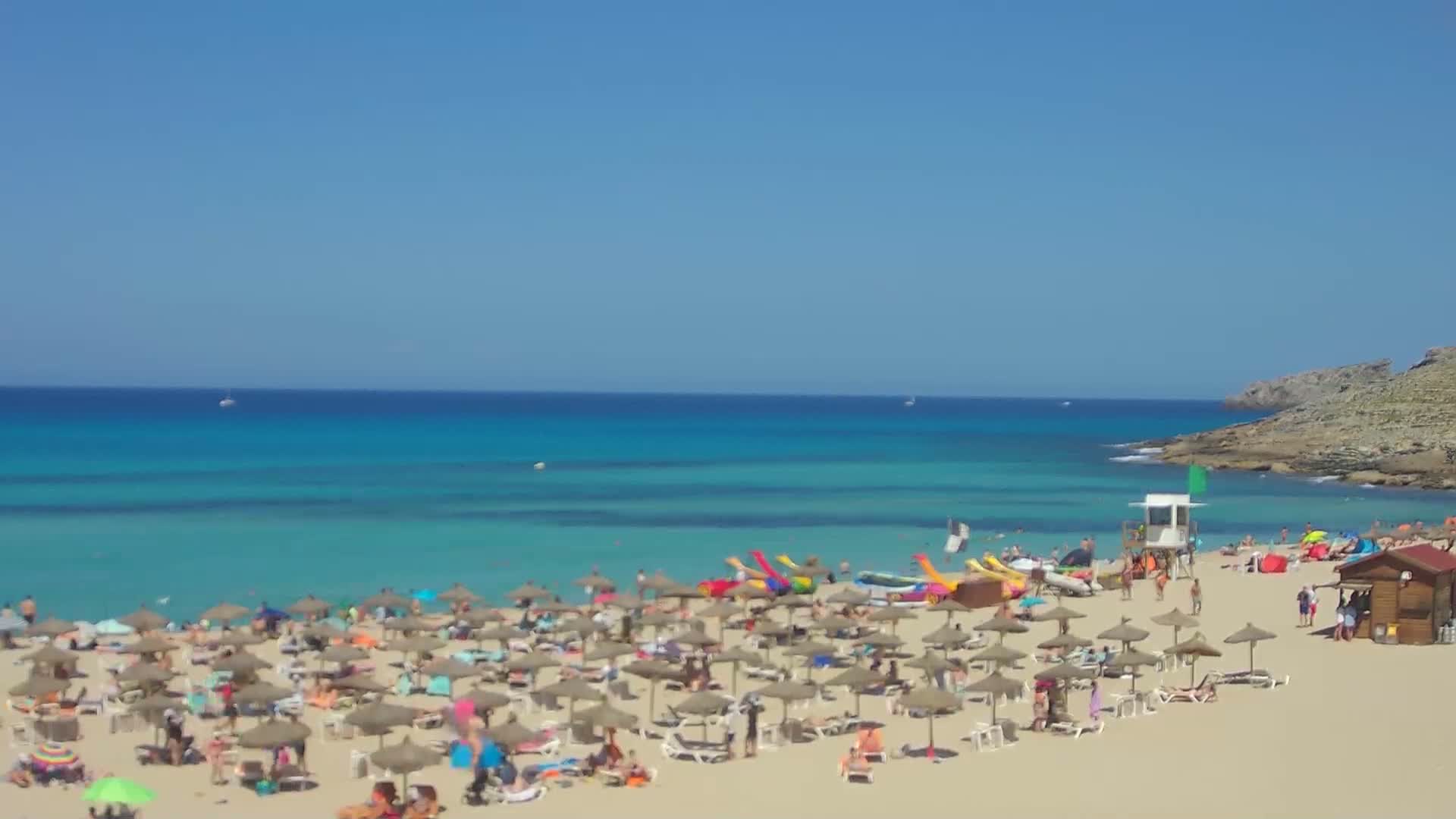 A sunny beach scene shows a sandy shore crowded with beach umbrellas and people, a lifeguard stand, and turquoise water extending to a clear blue sky.