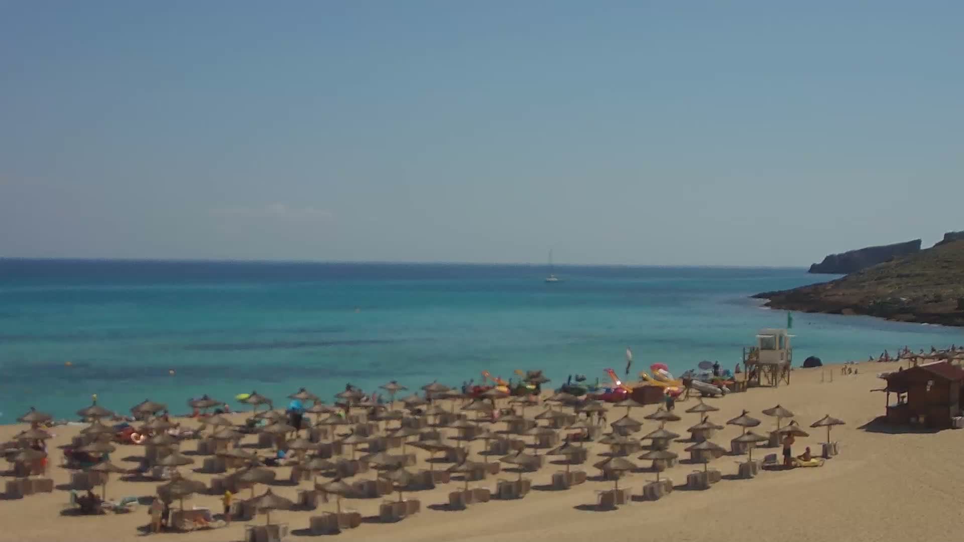 A sandy beach with numerous straw umbrellas and people under a clear blue sky, next to calm turquoise water and a small rocky landmass in the distance.