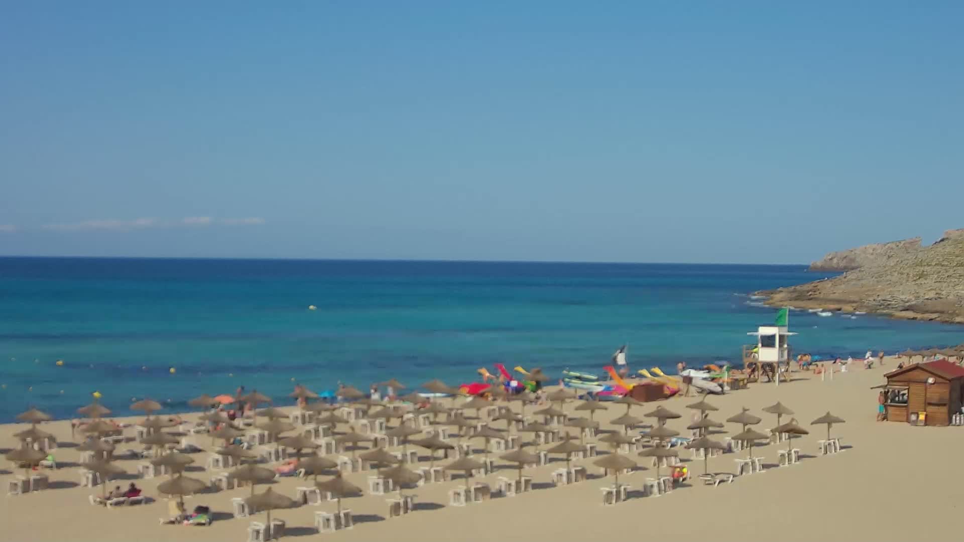A sandy beach with numerous straw umbrellas and lounge chairs sits beside a calm, turquoise ocean under a clear blue sky.