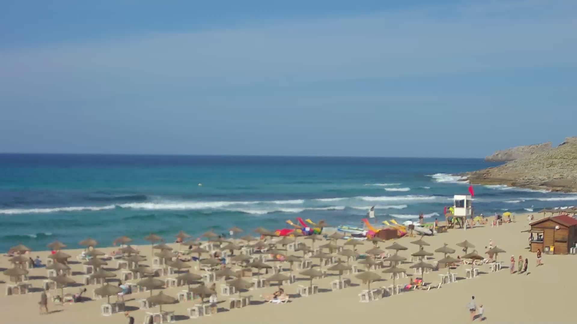 A sandy beach with numerous straw umbrellas and sunbathers is bordered by turquoise water with small waves under a clear blue sky.