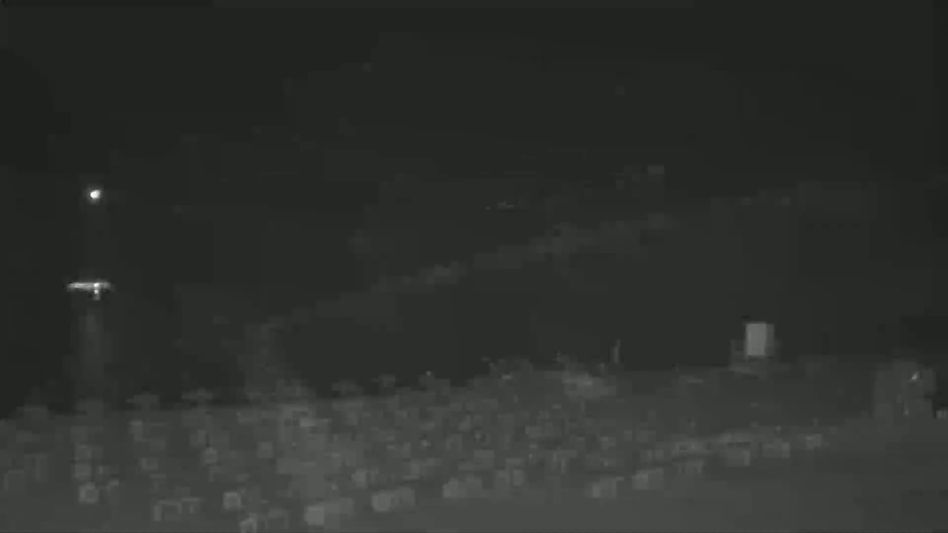 A nighttime, low-light image shows a large expanse of dark, indistinct objects, possibly vehicles or structures, with a tall, faintly lit structure in the left background.