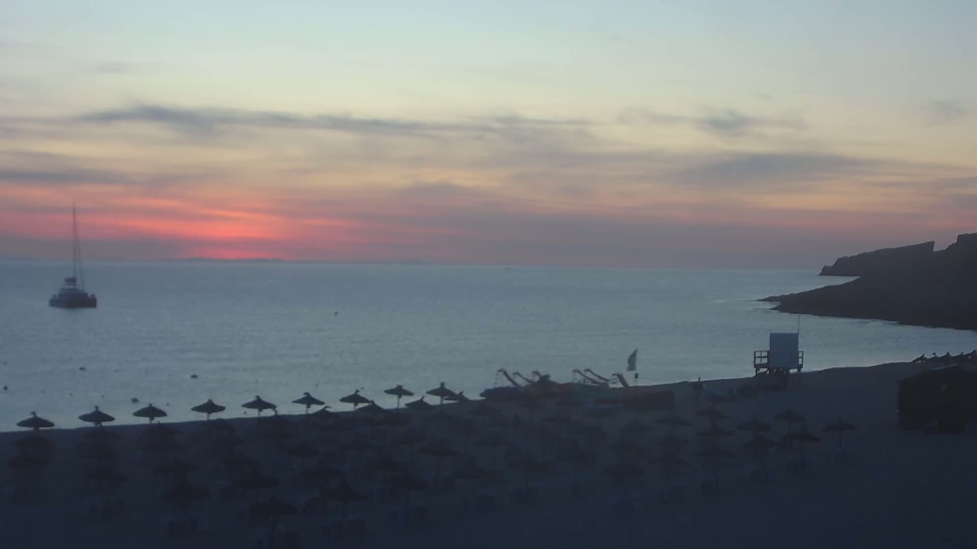 A calm sea at sunset with a boat, beach umbrellas, and a lifeguard stand on a sandy beach.