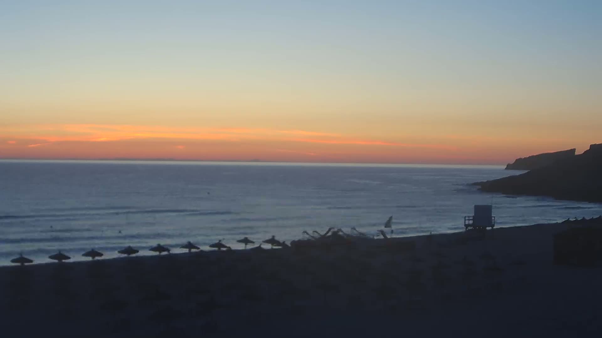 A beach at sunset with calm waves, a lifeguard stand, and a few closed umbrellas on the sand.
