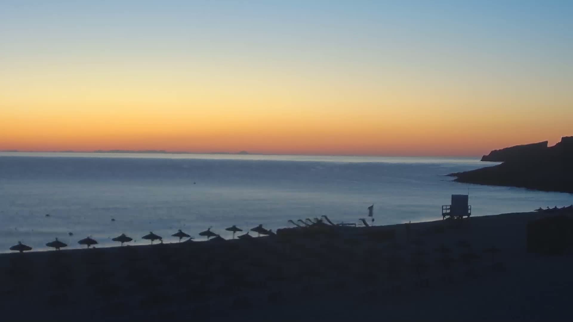A beach at sunset with several closed umbrellas and a lifeguard stand, situated next to a dark cliff overlooking a calm sea.