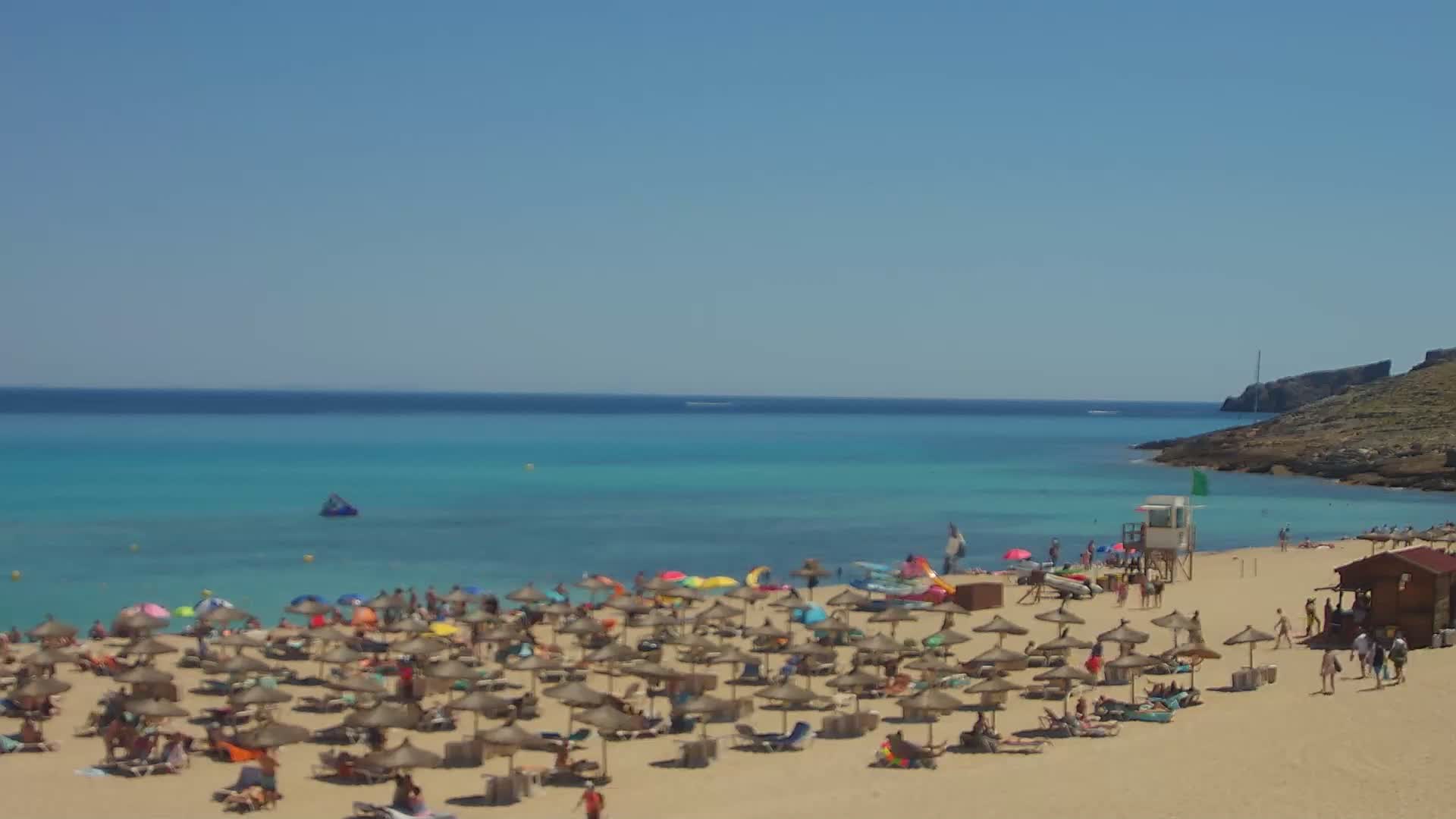 A sandy beach with many umbrellas and sunbathers is situated beside a calm, turquoise sea on a sunny day.