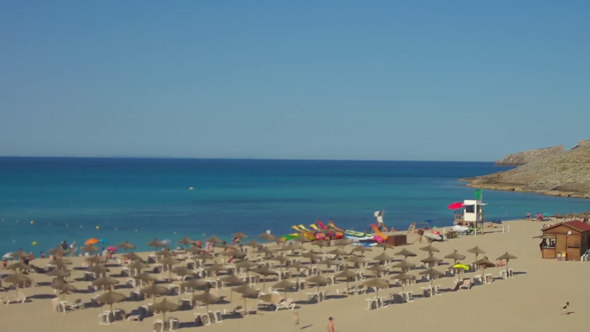 A sandy beach with numerous straw umbrellas and beachgoers is situated next to a calm, turquoise ocean under a clear blue sky.