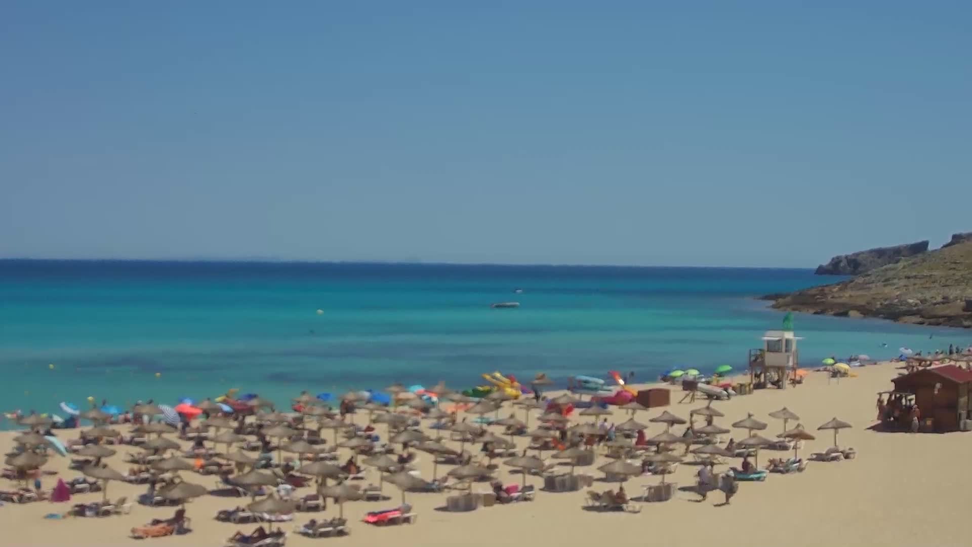 A sandy beach with numerous umbrellas and beachgoers is situated next to a calm, turquoise ocean under a clear, sunny sky.