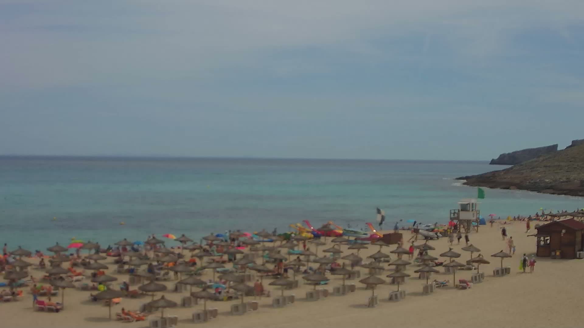 A sandy beach with numerous straw umbrellas and sunbathers stretches along a calm, turquoise sea under an overcast sky.