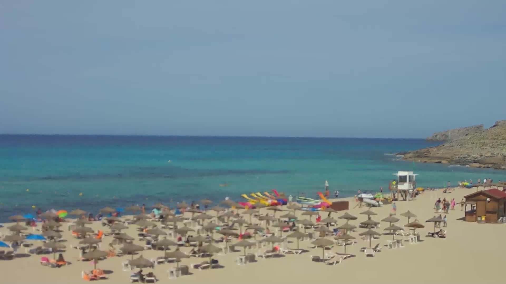 A sandy beach with many umbrellas and sunbathers stretches along a calm, turquoise sea under a clear sky.