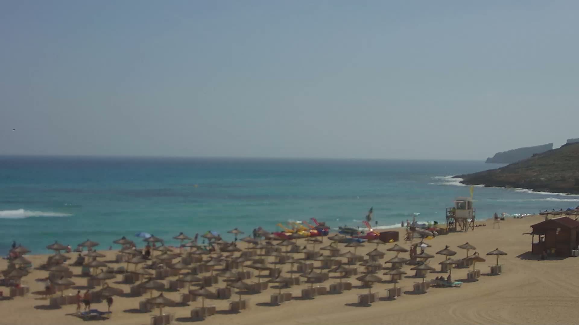 A sandy beach with numerous straw umbrellas, a lifeguard stand, and watercraft is seen under a clear, sunny sky next to a calm, turquoise ocean and a rocky headland in the distance.