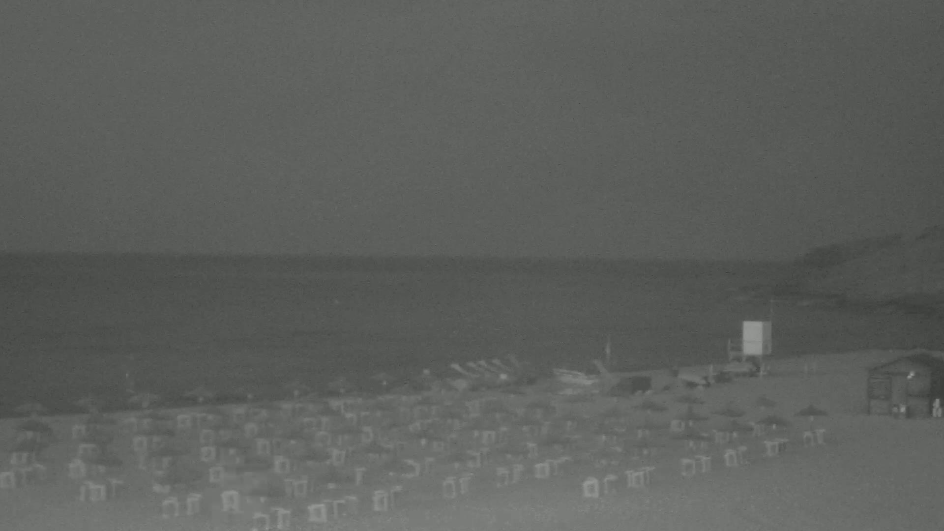 A beach at night under a dark sky shows many beach chairs and umbrellas, a calm sea, and a few structures on the sand.