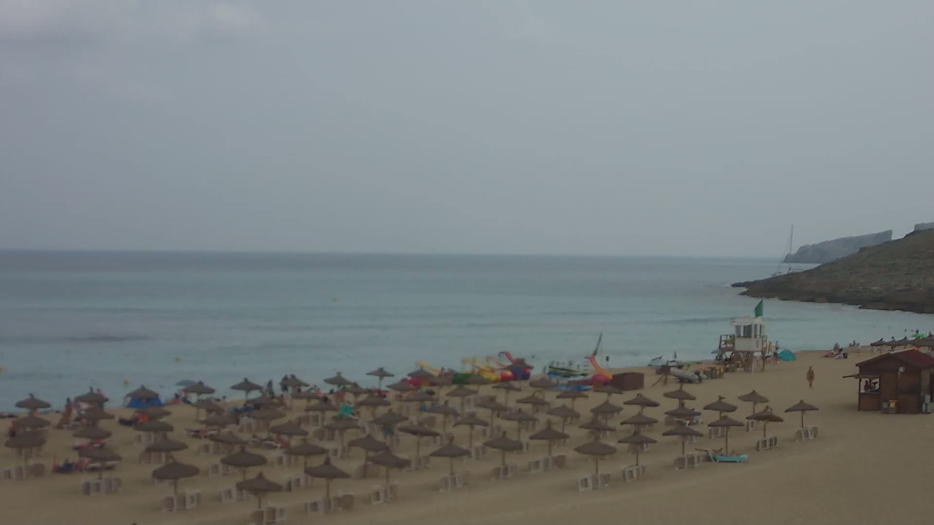 A sandy beach with numerous straw umbrellas and people, a lifeguard stand, and water toys, under an overcast sky, overlooks a calm, light-blue ocean.
