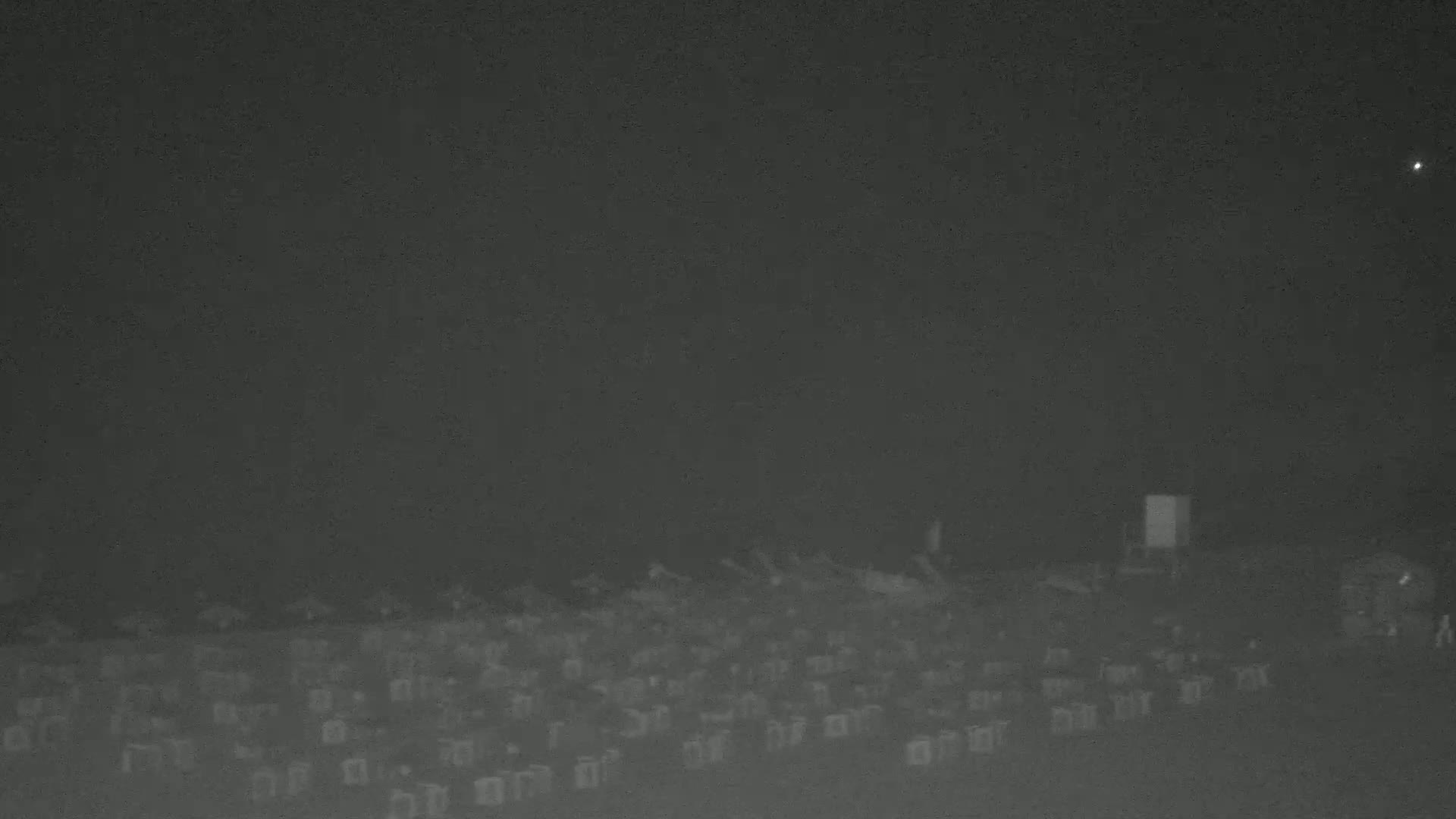 A low-light image shows a large, indistinct mass of objects, possibly chairs, arrayed in rows under a dark, hazy night sky.