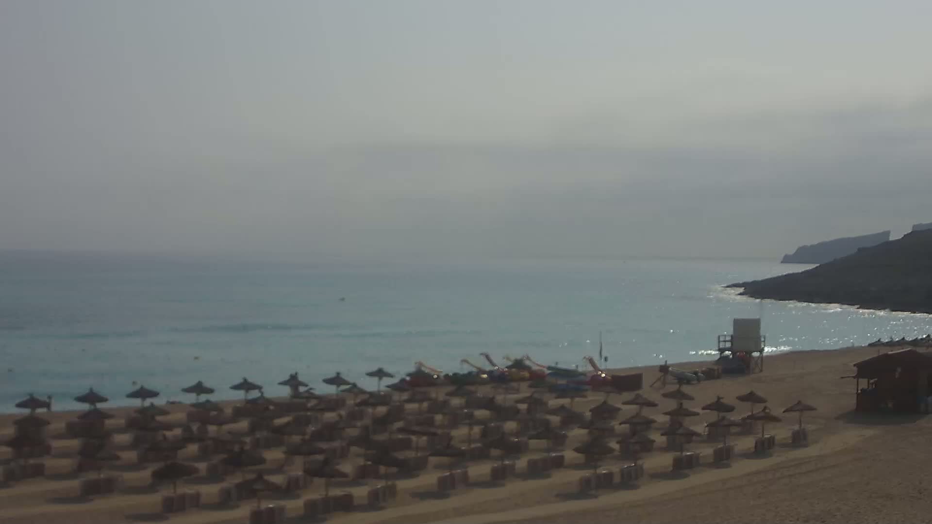 A hazy beach scene shows calm, light-blue water, a sandy shore lined with numerous straw umbrellas and beach equipment, a lifeguard stand, and a small building.