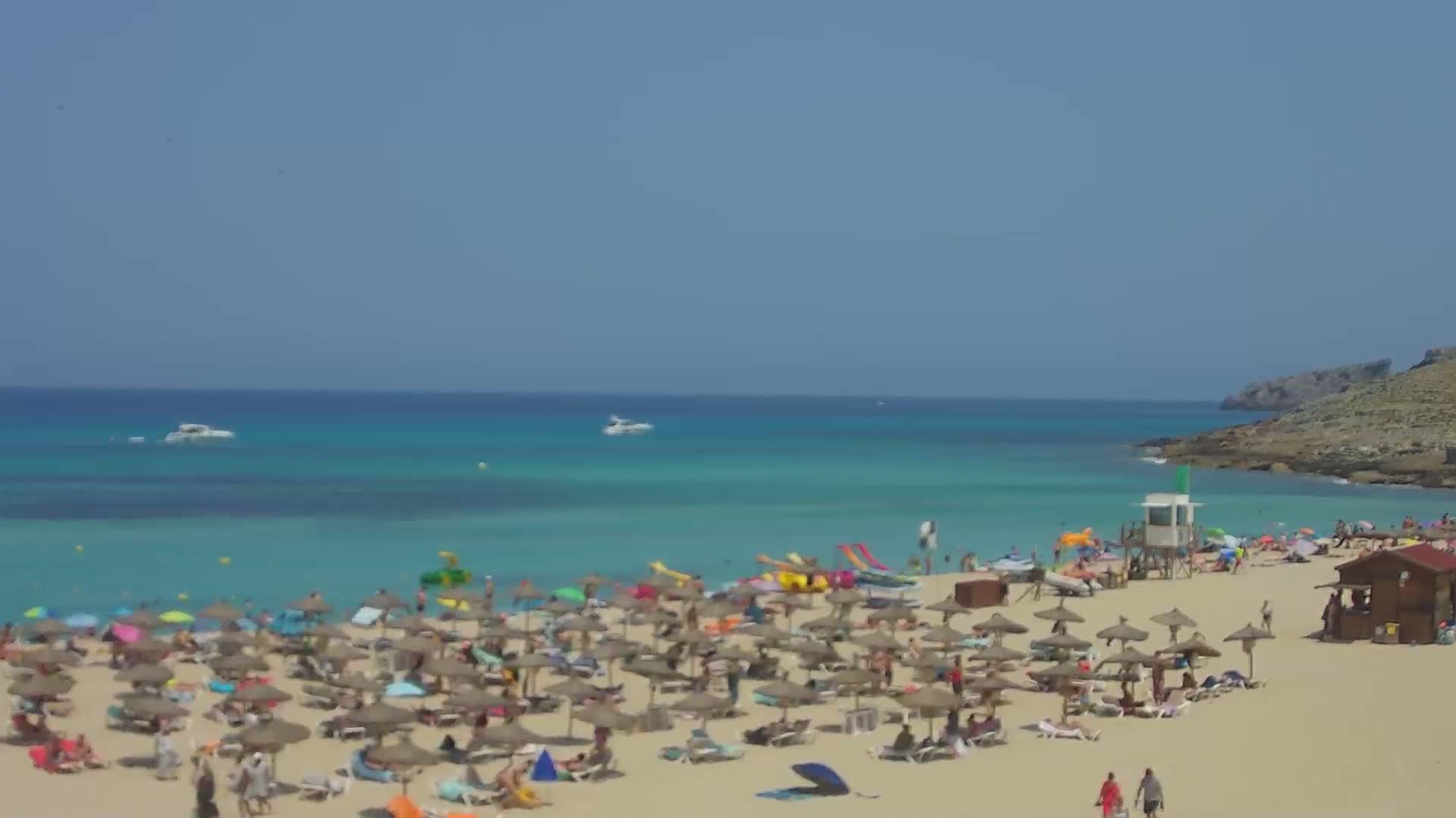 A crowded sandy beach with numerous umbrellas and sunbathers under a clear blue sky, features turquoise water and boats in the distance.