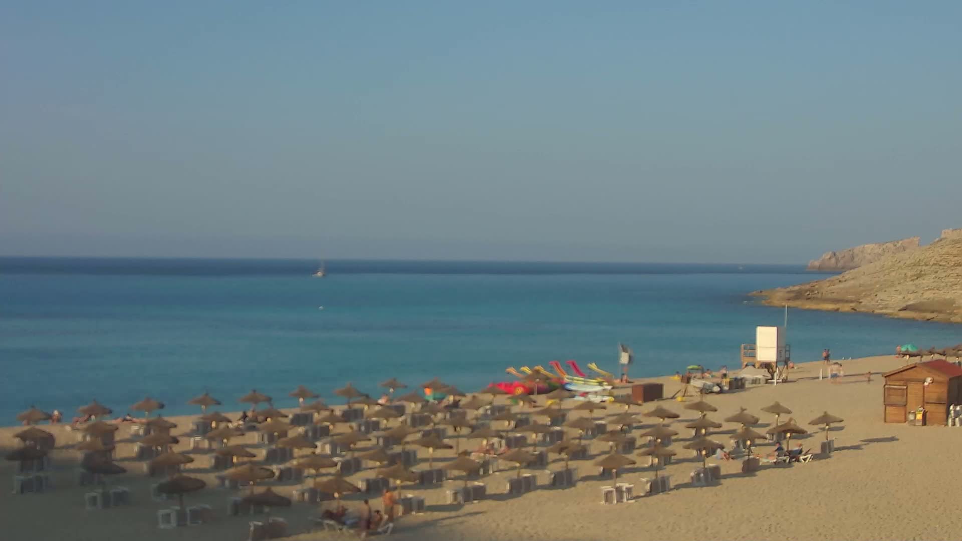 A sandy beach with numerous thatched umbrellas and sunbathers is situated on a clear, turquoise ocean next to a rocky cliff under a bright, sunny sky.