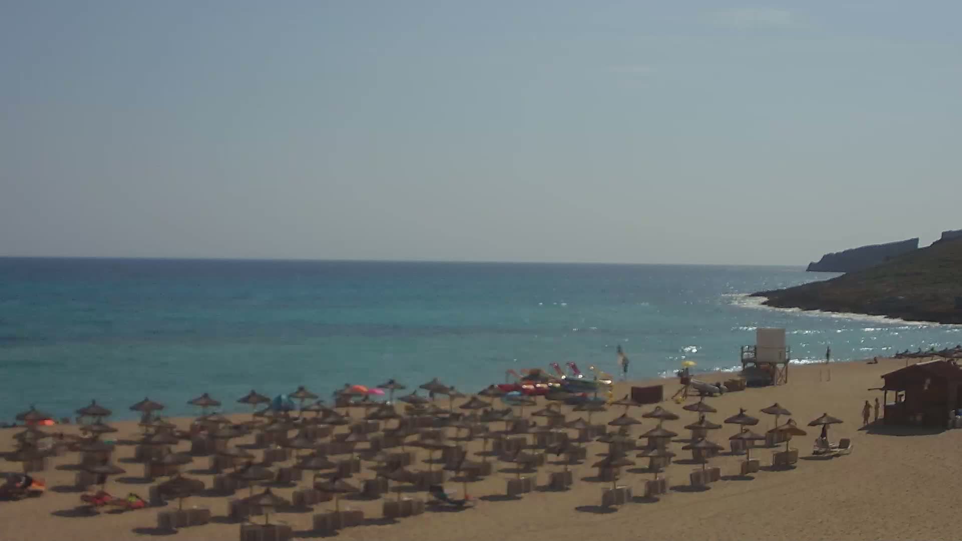 A sandy beach with numerous straw umbrellas and a lifeguard stand is situated on a calm, teal ocean under a clear sky.