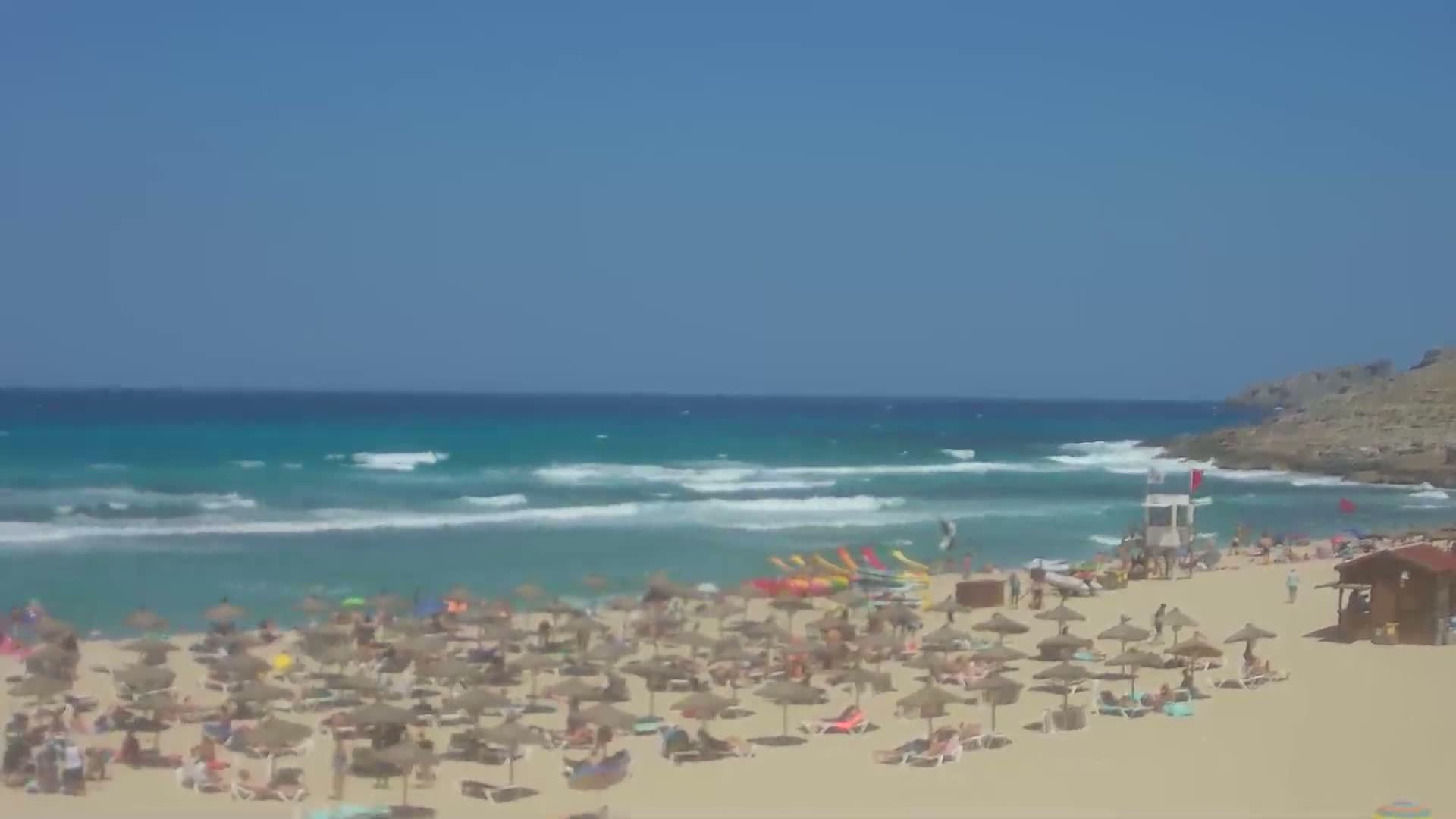A sandy beach with many people under umbrellas, and a lifeguard stand is visible near turquoise water with small waves under a clear blue sky.