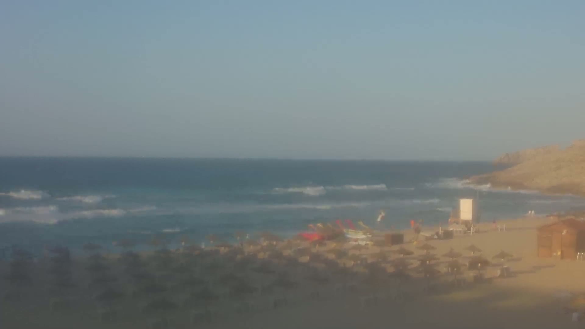 A hazy view of a beach with small waves, numerous beach umbrellas, and a few people under a mostly clear sky.