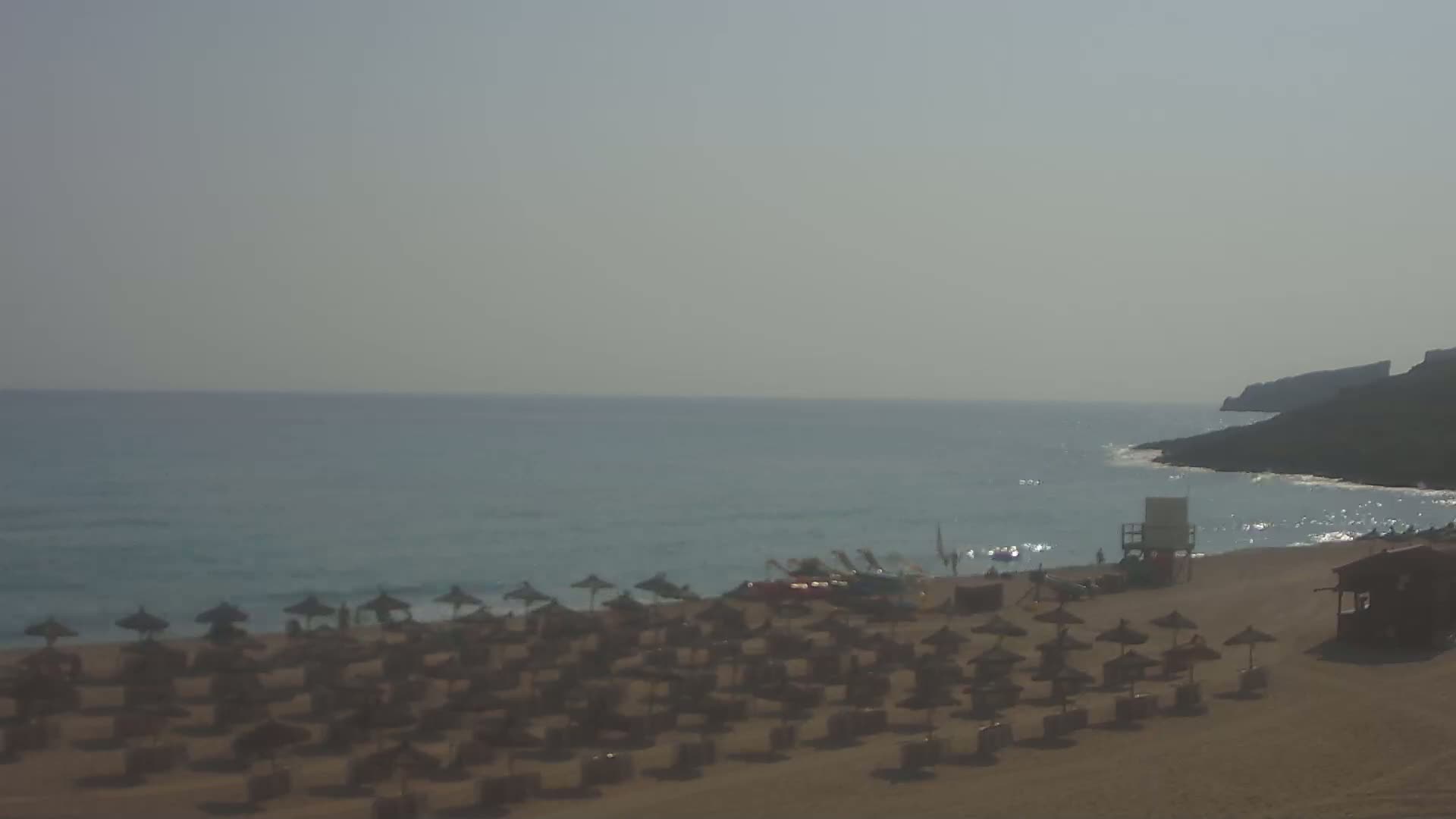 A sandy beach with numerous closed umbrellas and a lifeguard stand is situated beside a calm, light-blue ocean under a hazy sky.