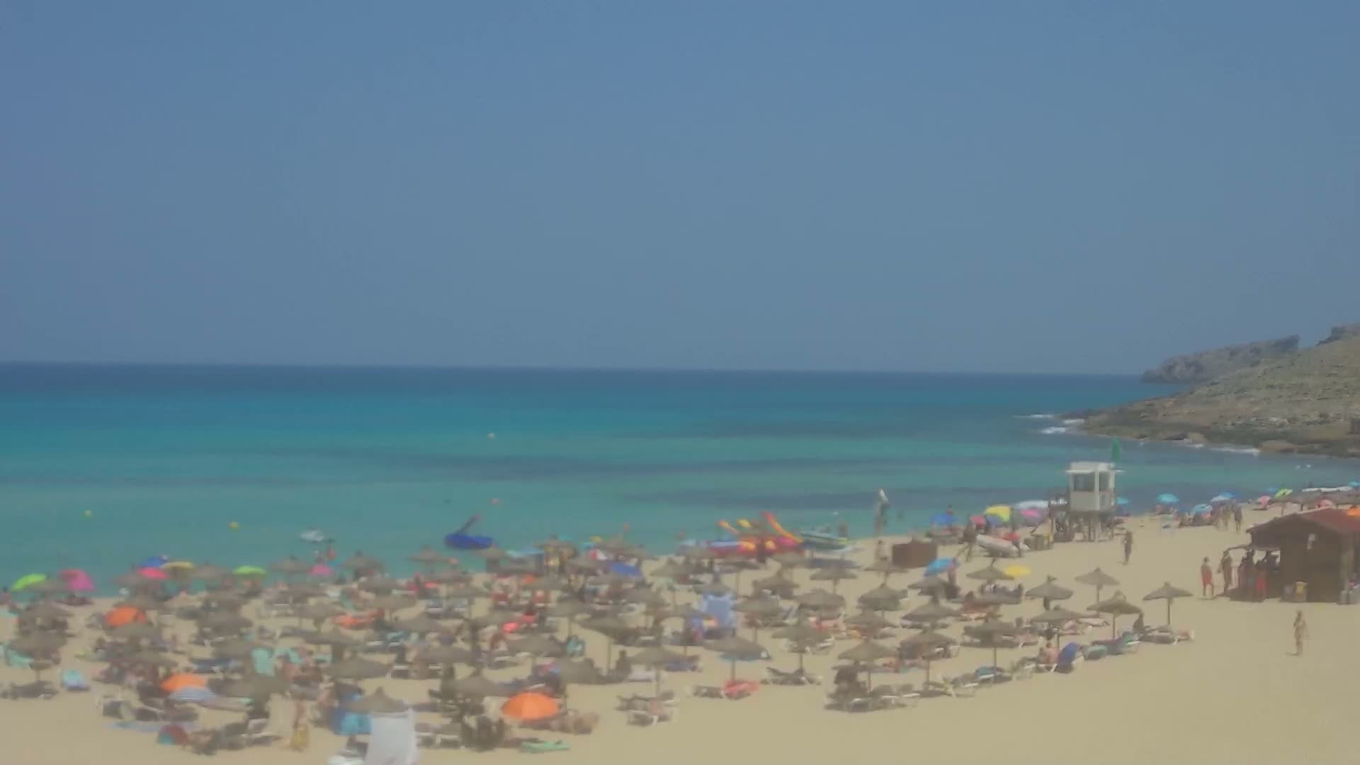 A crowded sandy beach with turquoise water under a clear blue sky features numerous beach umbrellas and sunbathers.