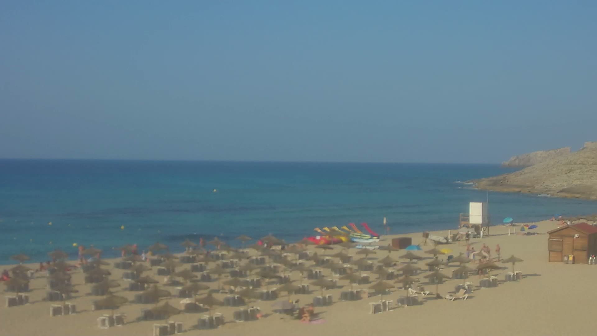 A sandy beach with numerous thatched umbrellas and people relaxing under a clear blue sky, next to a calm, turquoise sea.