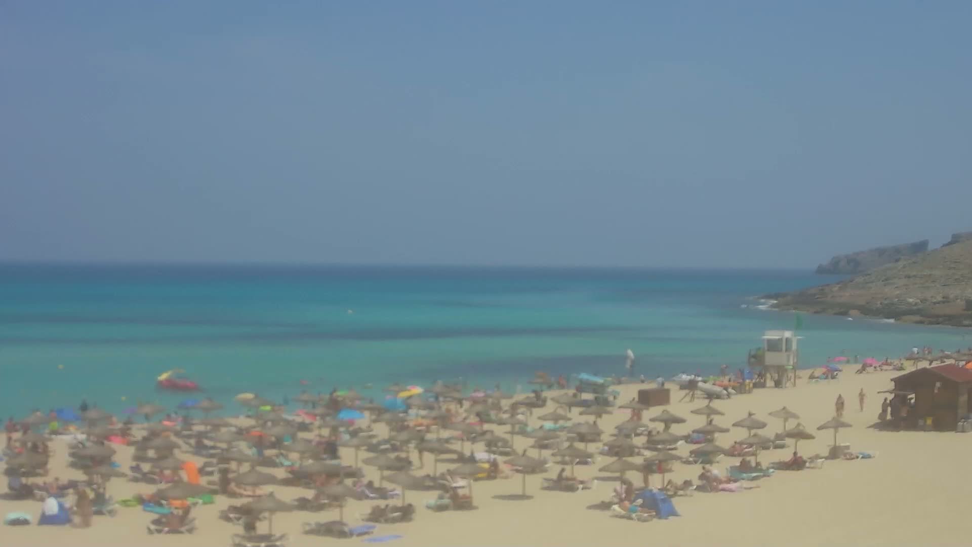 A sandy beach with numerous umbrellas and sunbathers is situated along a calm, turquoise ocean under a clear blue sky.