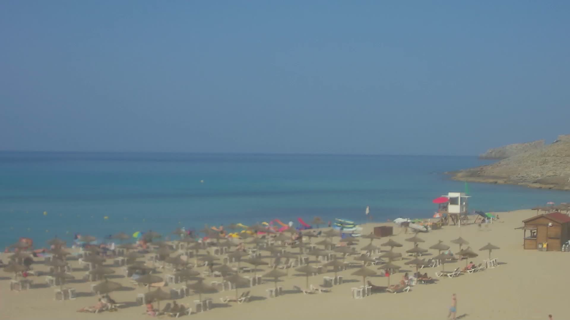 A sandy beach with numerous straw umbrellas and sunbathers stretches along a calm, turquoise sea under a clear blue sky.