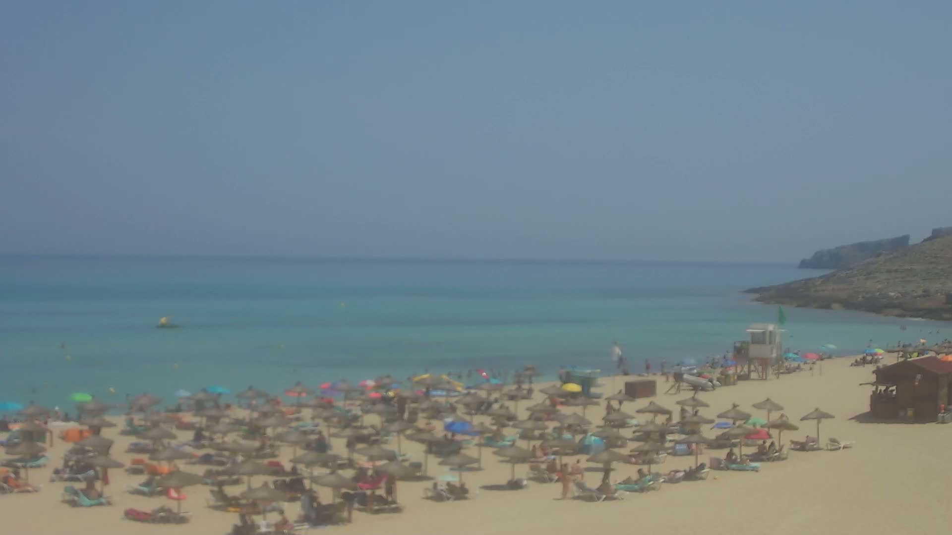 A sandy beach with numerous umbrellas and people relaxing is shown,  backed by a calm, turquoise sea and a rocky headland under a clear sky.