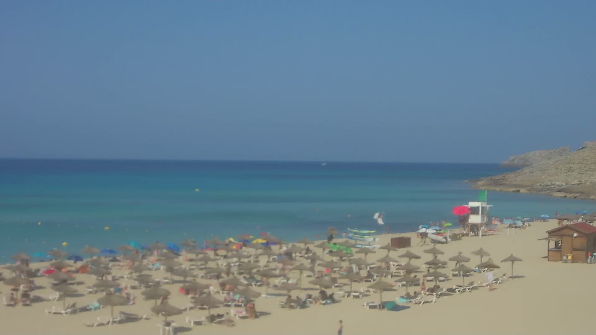 A sandy beach with numerous umbrellas and sunbathers is situated next to a calm, turquoise ocean under a clear blue sky.