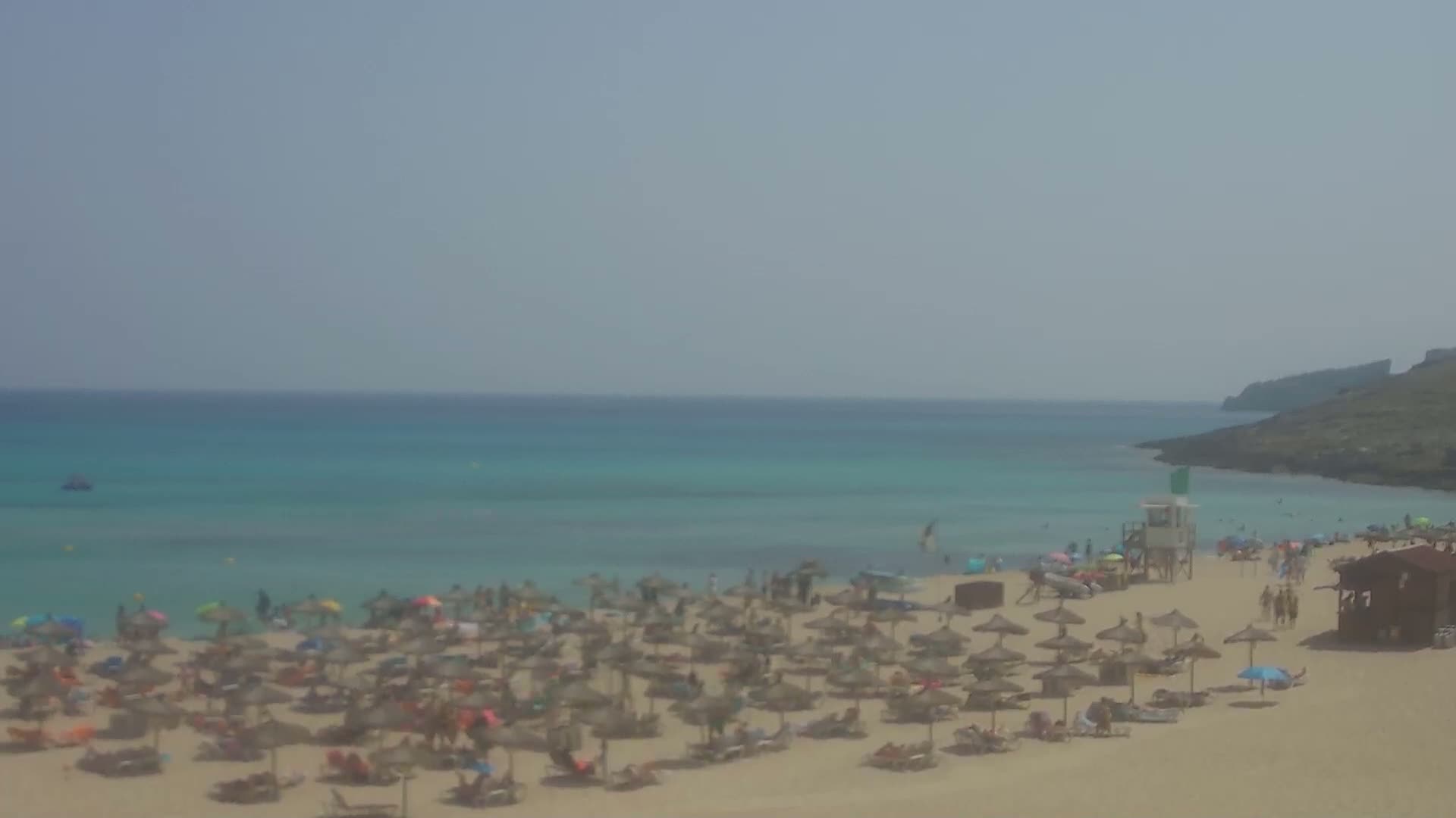 A sandy beach with many umbrellas and people, a lifeguard stand, and a calm, turquoise ocean under a hazy sky.