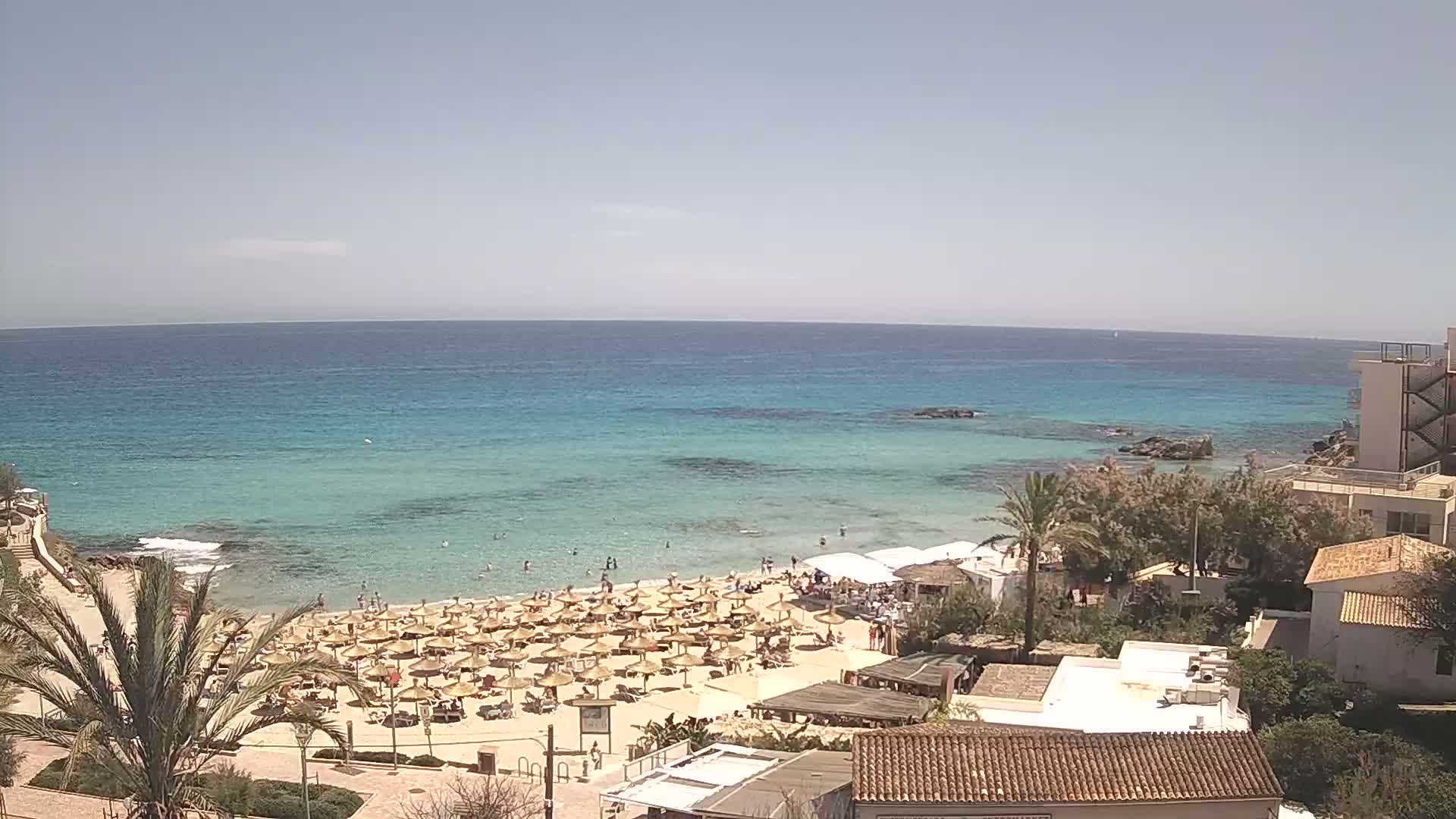 A sandy beach with numerous umbrellas and sunbathers is situated next to a calm, turquoise ocean under a clear, sunny sky.