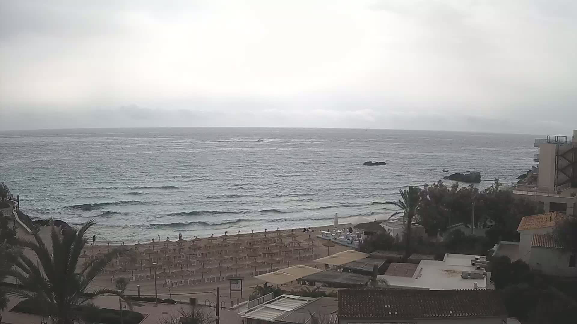 An overcast day shows a beach with many closed umbrellas, a calm sea with small waves, and buildings overlooking the water.