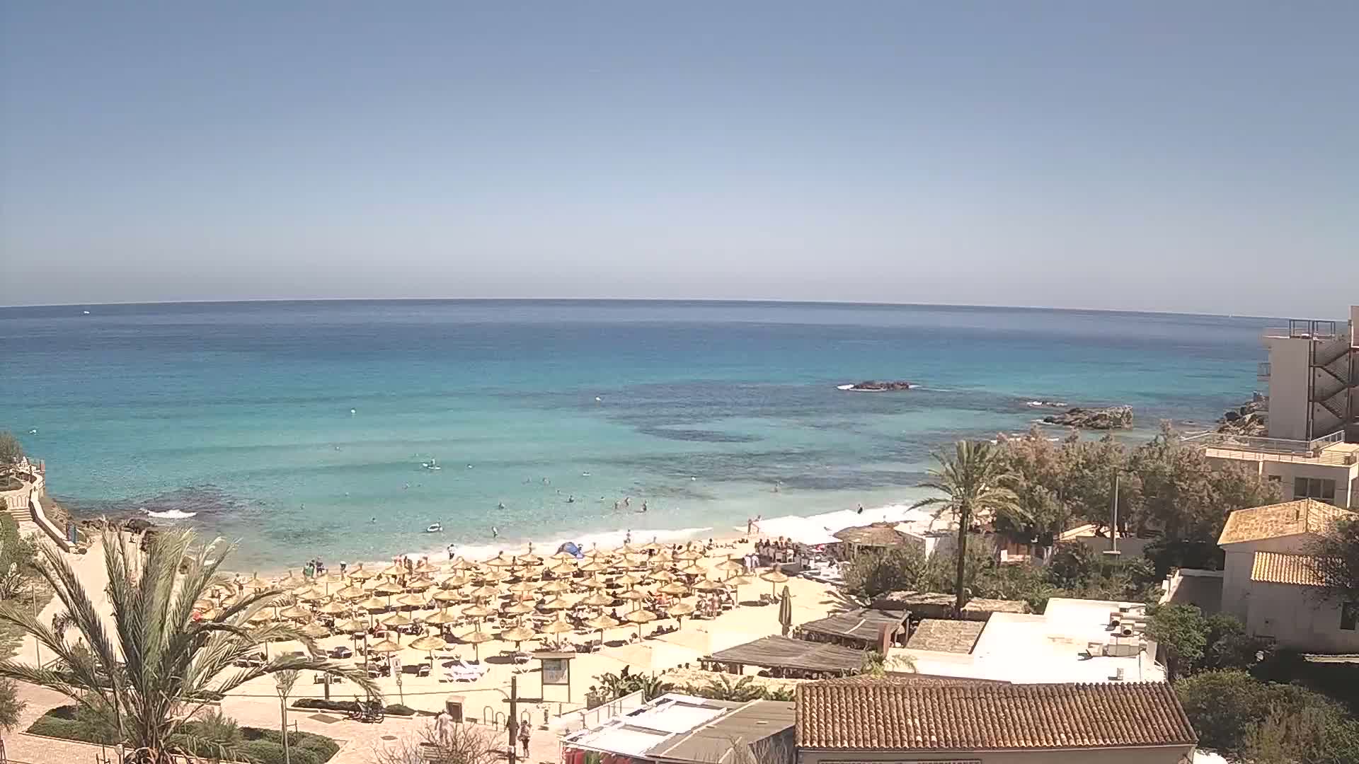 A sandy beach with numerous straw umbrellas is crowded with people, overlooking a calm, turquoise ocean under a clear blue sky.