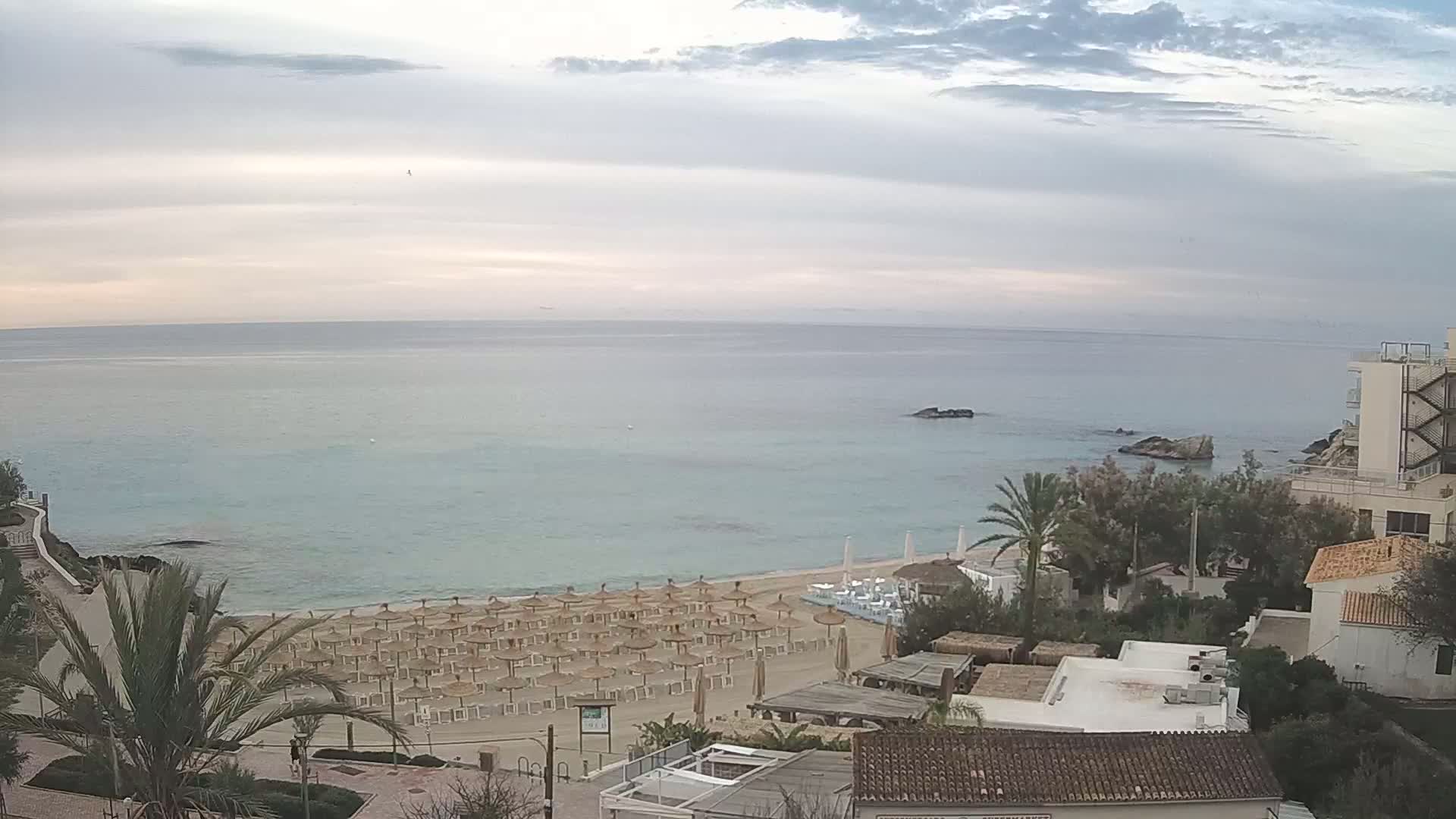 A calm, overcast day reveals a beach with numerous closed umbrellas and a few buildings overlooking a tranquil turquoise sea.