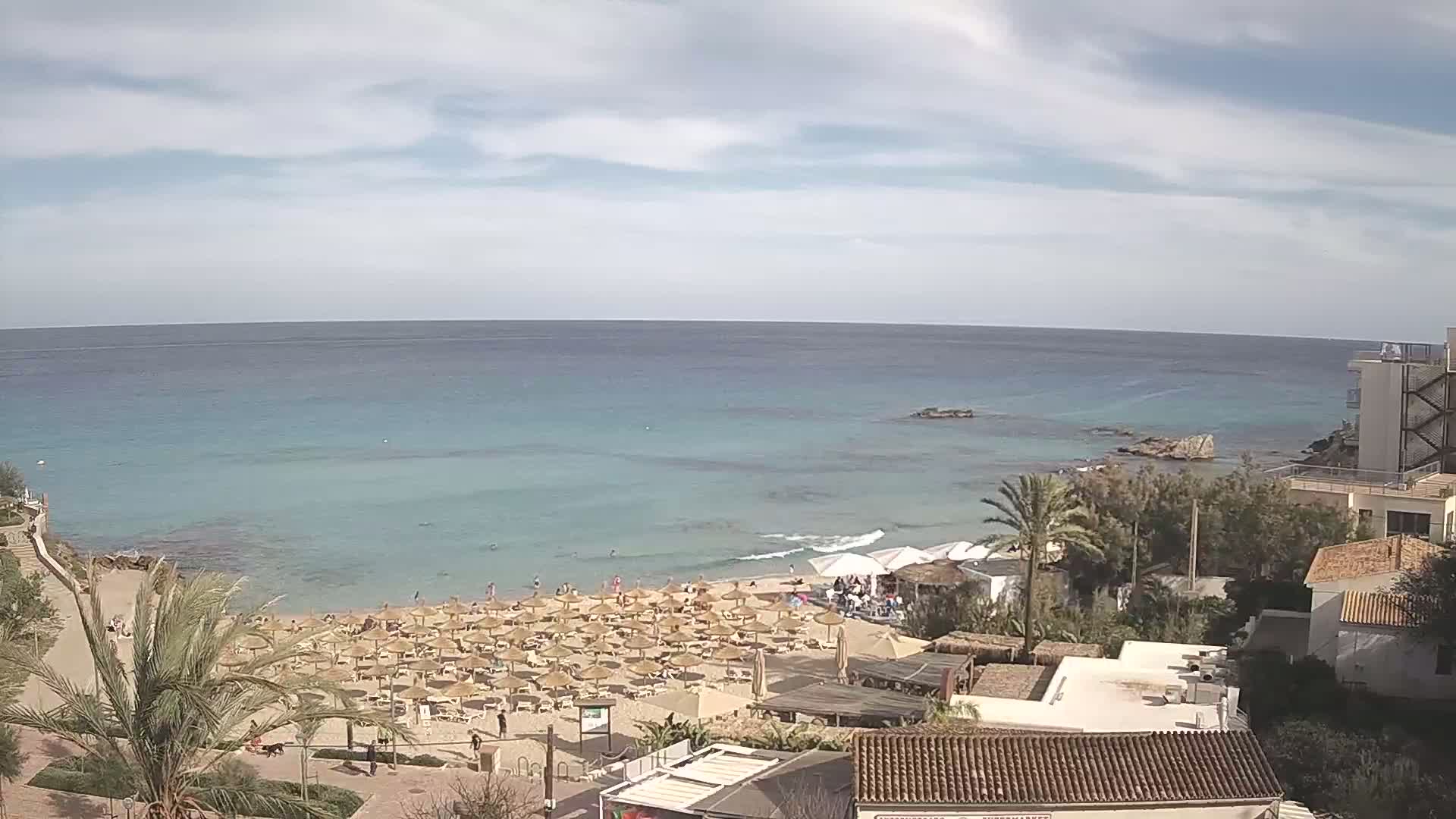 An aerial view shows a sandy beach with numerous straw umbrellas under a partly cloudy sky, next to a calm, turquoise sea and buildings.