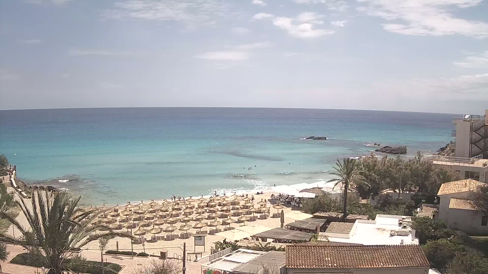A sandy beach with numerous straw umbrellas and a calm, turquoise sea under a partly cloudy sky is overlooked by buildings and palm trees.