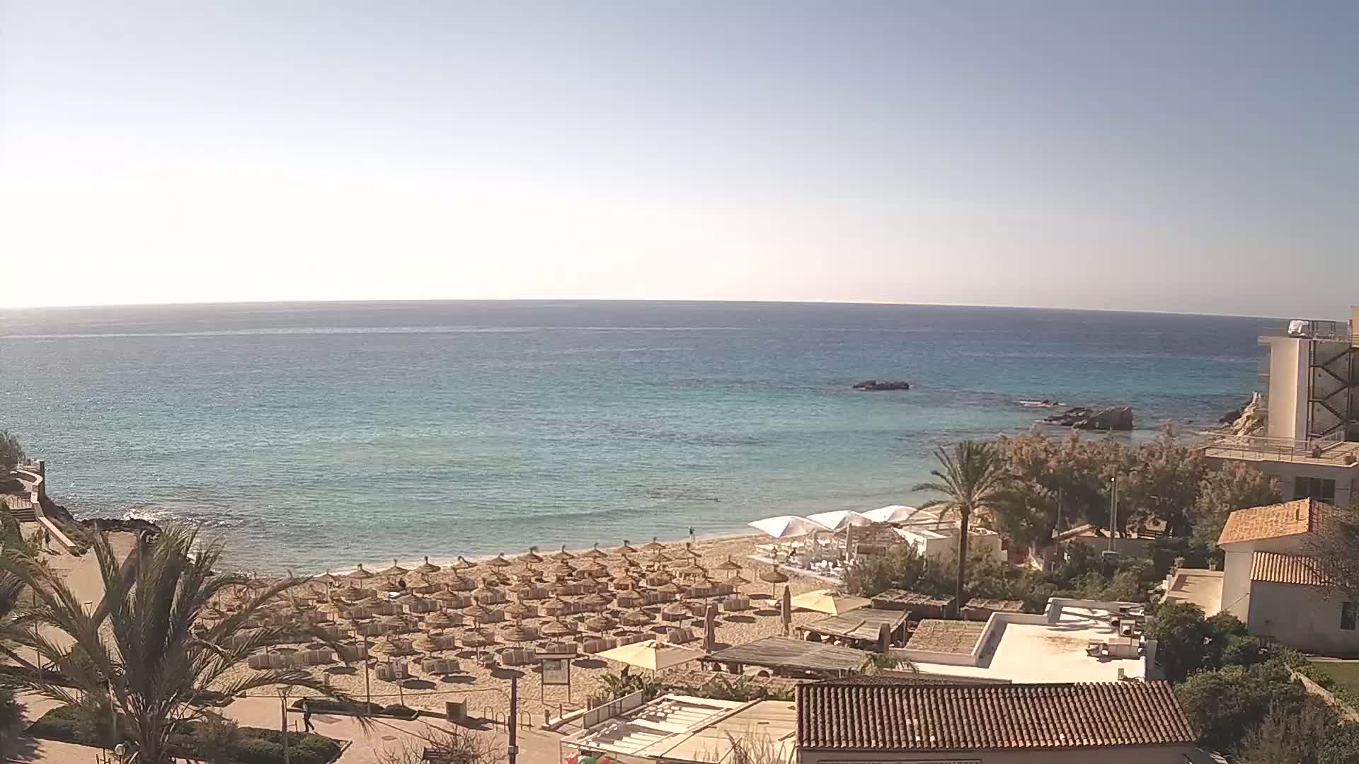 A sandy beach with numerous umbrellas and beach chairs overlooks a calm, clear turquoise sea on a sunny day.